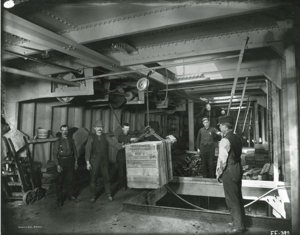 Factory workers are guiding a crated McCormick reaper on pulley ropes through an opening in the floor, most likely at the McCormick Reaper Works. The text on the wooden crate reads: "McCormick Folding Daisy Reaper. Chicago, U.S.A. D. No.2. Gross 291 Kilo. Net 231 Kilo. McC. Hamburg."