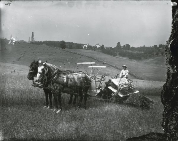A man is using a McCormick grain binder pulled by two horses to work in a field on a hillside. The hill overlooks bundles of the harvested crop. In the background are a horse and carriage tied to a fence, and farm buildings. In the distance on the left are what appear to be five large derricks.