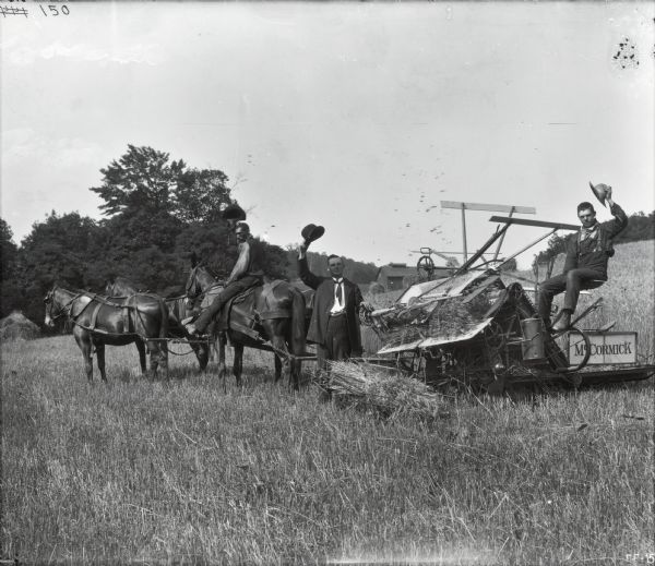 Two men are lifting their hats and posing with a McCormick grain binder in a farm field. Another man is lifting his hat while sitting on one of four horses pulling the binder.