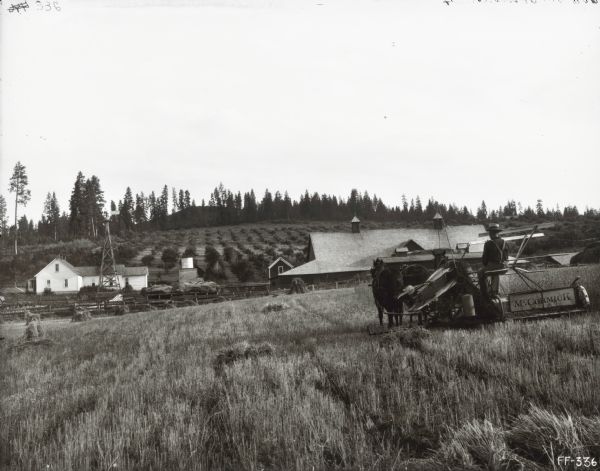 A man is using a McCormick grain binder pulled by horses to work in a field. In the background is a barn, windmill and several other farm buildings.