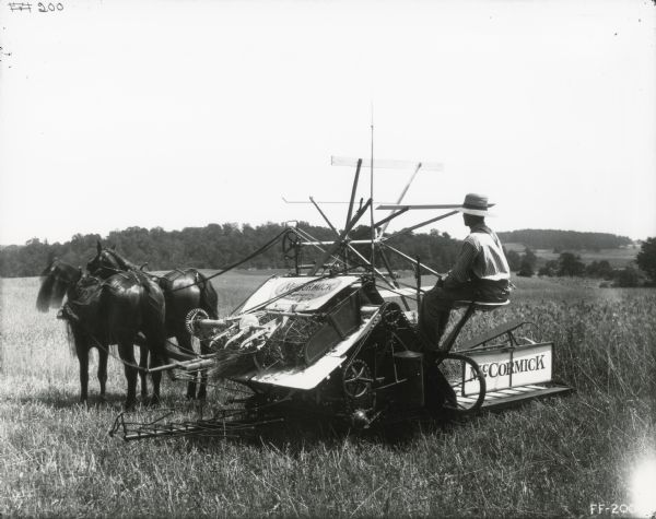 A man wearing a hat, work clothes and suspenders is operating a McCormick grain binder in a farm field.