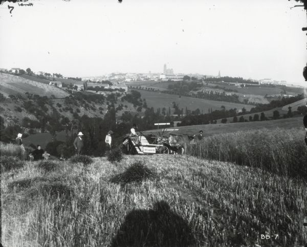 Men use a McCormick grain binder to harvest a crop on a hill overlooking a valley. Town buildings are on a hill in the far background. Two women and two children are on the left behind the binder. The photographer's shadow is in the foreground.