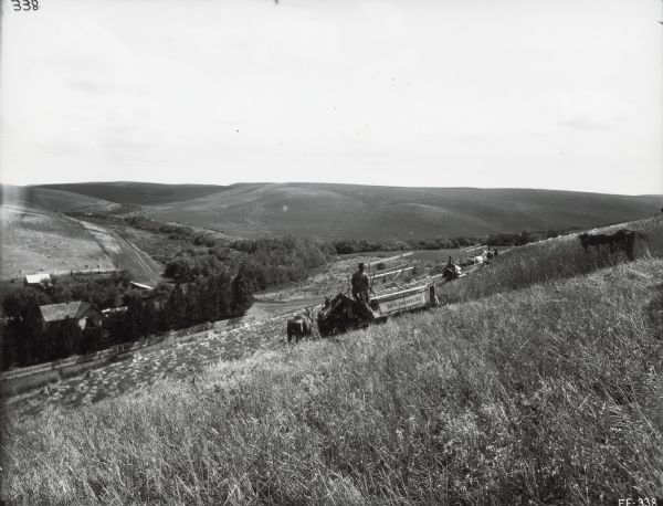 Two farmers are using McCormick grain binders along the side of a steep hill overlooking farm buildings below.