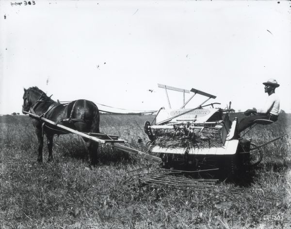 Side view of a man operating a horse-drawn McCormick twine grain binder in a field.
