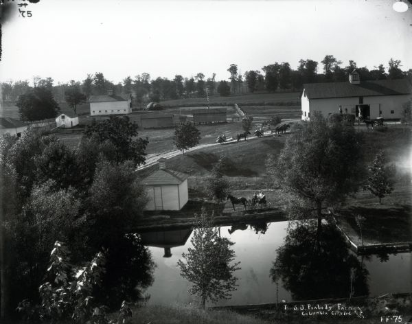Elevated view of multiple buildings and a body of water on the farm of S.J. Peabody. Three men are riding in a horse-drawn carriage along the water in the foreground, while two men are sitting on two McCormick binders pulled by horses on a drive leading to a barn. Other people are posing near the barn, on the grass and near a flagpole.