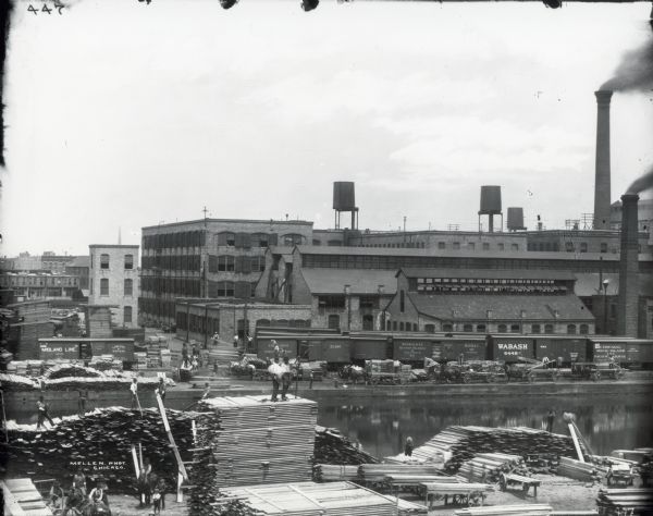 View across canal of the McCormick Reaper Works factory, rail yard, and surrounding buildings. Men in the foreground are stacking piles of lumber. After 1902, the factory became International Harvester's McCormick Works.
