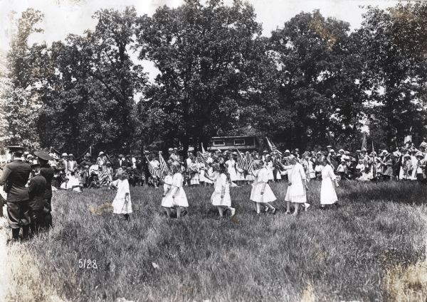 A group of girls performing a flag drill during a School Festival as a crowd of adults and children look on. Three boys are standing on the left playing musical instruments.