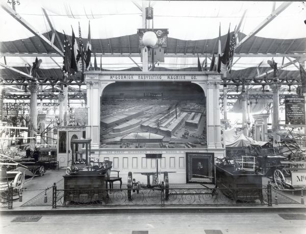 Flags hanging above a McCormick Harvesting Machine display at what is possibly the World's Columbian Exposition (The World's Fair of 1893). A painting of a bird's-eye view of the McCormick Reaper Works factory is hanging as the centerpiece to the display, and a framed portrait of Cyrus Hall McCormick, set on the ground, is leaning against it.