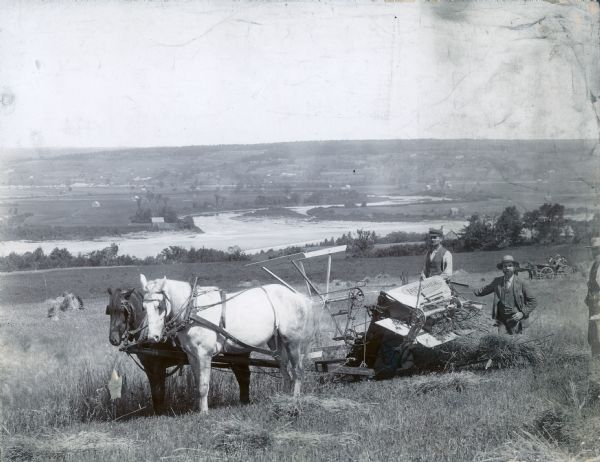 Three men use a McCormick grain binder in a field overlooking a body of water, probably the Saint John River, in Fredericton, New Brunswick, Canada. Men and women riding in a horse-drawn carriage are in the middle background.