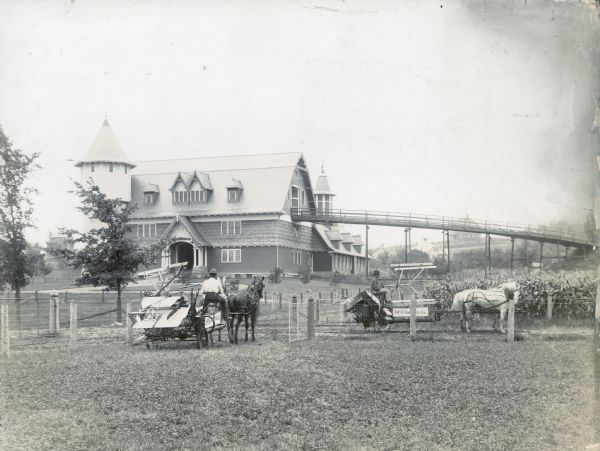 Two men use horse-drawn McCormick grain binders in a field in front of a dairy barn at the University of Wisconsin-Madison. A grain elevator or walkway extends from the barn's loft.