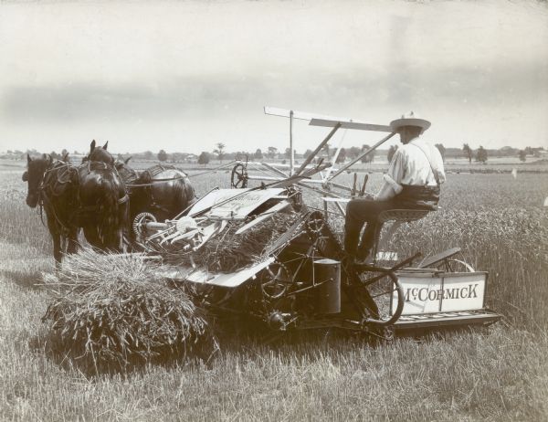 A man drives two horses in pulling a McCormick binder across a field while completing farm work.