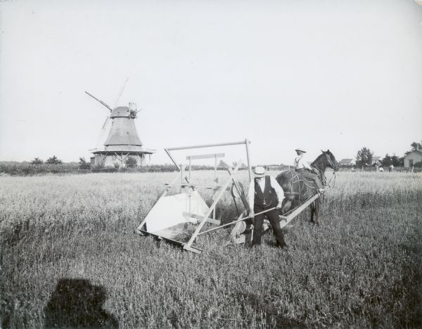 A man wearing a hat and smoking a pipe is raking cut stalks from a binder as a boy is riding a horse pulling the machine across a field. A windmill is standing in the background on the left. The photographer's shadow is visible in the foreground.