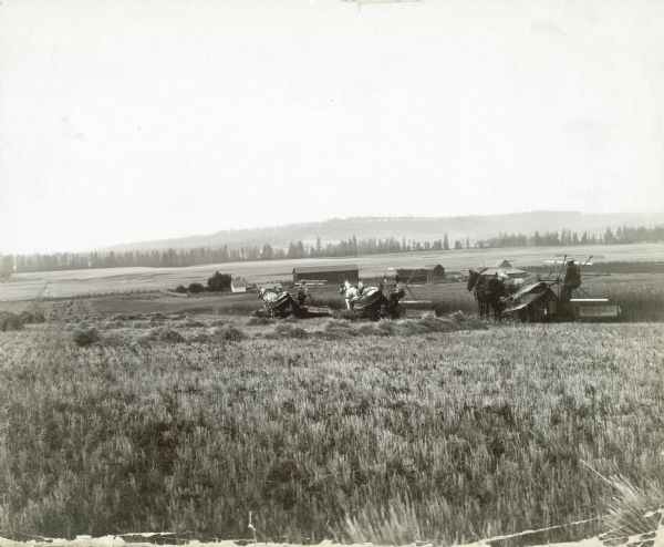 View from a distance of three farmers, and perhaps a child, using horse-drawn McCormick grain binders in a field. Farm buildings are in the middle distance.