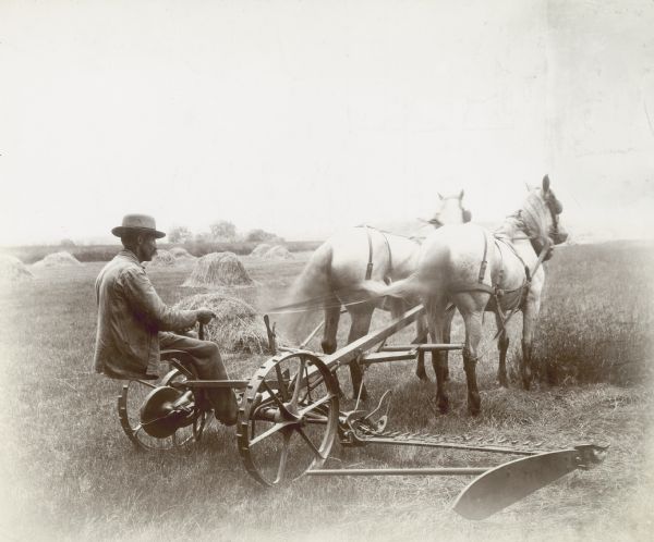 A man is using two horses to pull a McCormick mower through a field, probably in or around Boston. Piles of what appear to be hay are in the background.