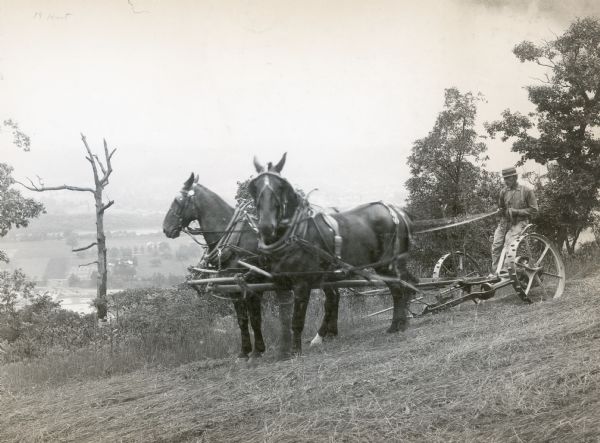 A man is using a 7-foot McCormick mower on hill on the farm of William Knoderer, overlooking what appears to be a body of water and farm buildings.