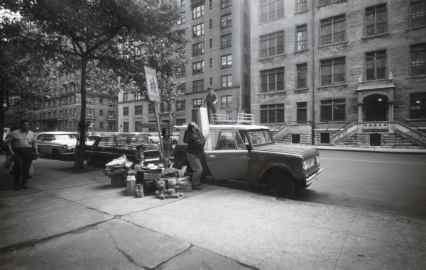 Shelly and Mary Louise Grossman, and their children Keith and Julie, load items including teddy bears, suitcases, and boxes into an International Scout truck parked along the side of an urban street.