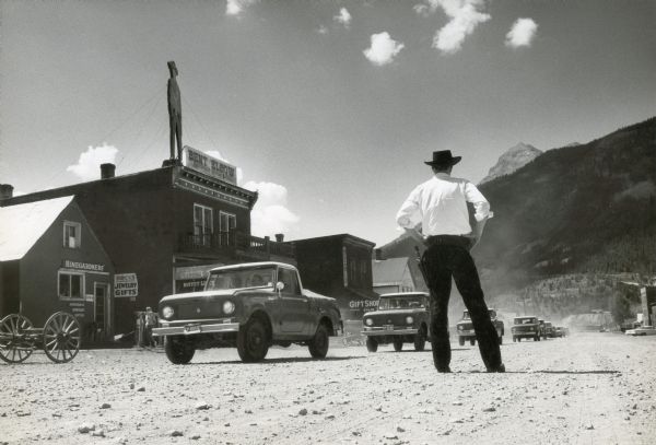 A man wearing a cowboy hat and a hip holster stands on a dirt road as a line of International Scout trucks passes by. A series of shops, restaurants, and other buildings lines the other side of the road (probably Blair Street).