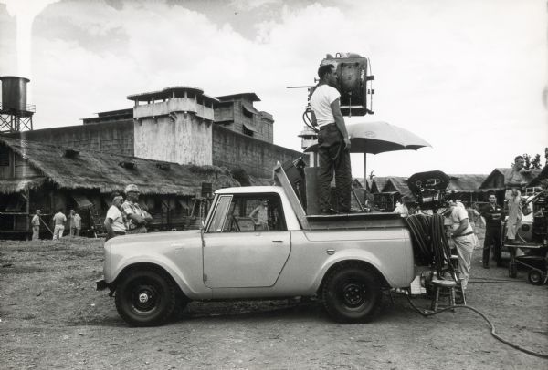A man stands on the bed of an International Scout truck while using a stage light on the set of the film "King Rat." Thatched-roof huts, a water tower, and cement buildings are in the background, and other men stand near the truck.