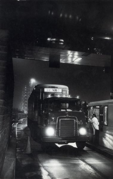 A man leans out of the tollbooth on George Washington Bridge to accept payment from the driver of an International semi-truck marked, "Gomez." George Washington Bridge spans the Hudson River, connecting the borough of Manhattan in New York City to Fort Lee, New Jersey.