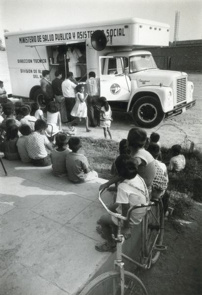 A group of children gathers around an International truck modified for use by a public health organization while a woman in a nurse's outfit stands inside. The words on the truck, written in Spanish, read, "Ministerio de Salud Publica u Asistencia Social. Direccion Tecnica..."