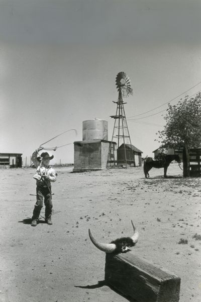 6-year-old Kirk Hanna uses a lasso to practice cattle roping on a pair of bull horns affixed to a log in a barnyard. A horse is tied to a fence on the right, and a windmill and other farm buildings are in the background.