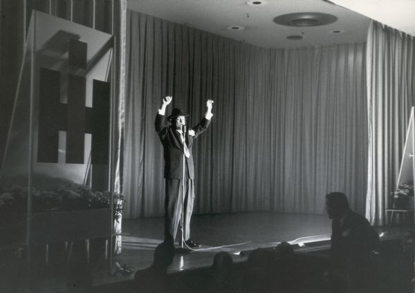 Red Skelton entertains an audience while onstage at the convention of American Trucking Associations. The International Harvester logo is displayed on the left of the stage.