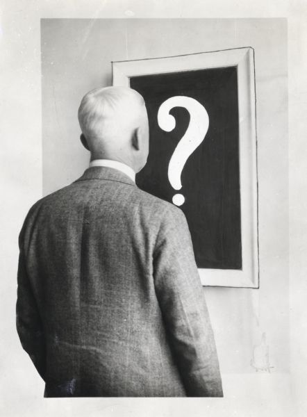 F.J. Wolfe stands with his back toward the camera while looking at a framed picture of a question mark hanging on a wall. The original typewritten caption reads: "Title: Portraits. Back view of F.J. Wolfe taken for the Boys and Girls Book."  