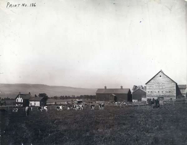 A man wearing overalls, a man with a dog, and a man driving a team of two horses are posing among a herd of cows on a farm. Several barns, a fence, another herd of cows, and a farmhouse are behind the men. In the far background are hills.