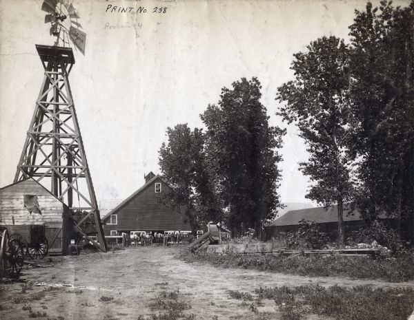 View down dirt road of farm buildings and a windmill. In the background in front of a barn men stand with horses near a fence.