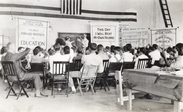 A group of men and women sit in chairs inside what appears to be a classroom or gymnasium while practicing knot-tying.  They follow an instructor's example as he stands near several posters set up near a wall.
