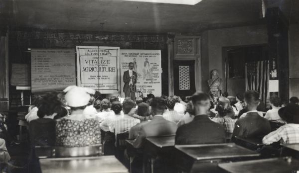View from back of classroom of a group of teachers sitting at desks in a classroom while observing an instructor demonstrate knot tying. Several posters hang on the wall behind the instructor.