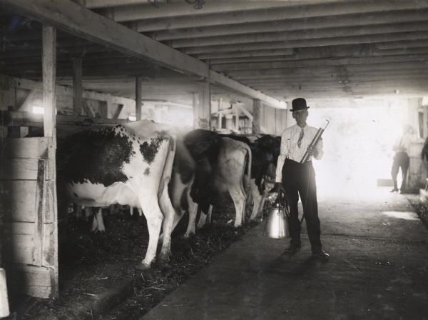 A man wearing a bowler hat, tie, and suspenders is holding milking equipment while standing near a row of cows inside a barn. Another man is standing in the background near the open barn door.