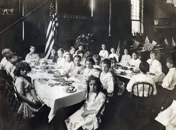 A group of children sitting around tables at the Second Grade Normal School. The room is decorated with flowers and American flags.