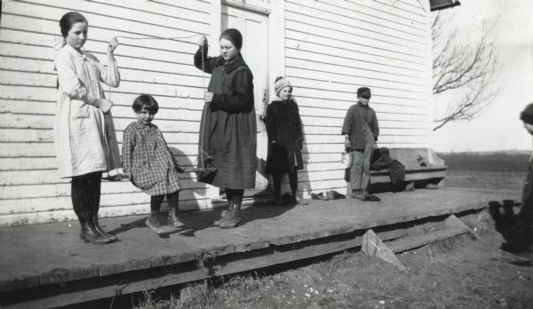 Two girls standing on the wooden porch of a schoolhouse holding ropes attached with timber hitch knots to a piece of wood on which a girl is sitting. Other children are standing on the porch and on the ground nearby.