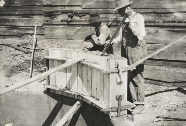 Two men wearing hats pouring cement from a bucket into a wooden form to build a foundation for a silo. Behind them is a farm building.