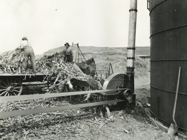 Two men are unloading silage from a wagon into a silo filler on the farm of Adolph Eichherst. The silo filler is hooked up to a belt for power. Original caption reads: "Outfit owned by Reed Roy, Wheaton."