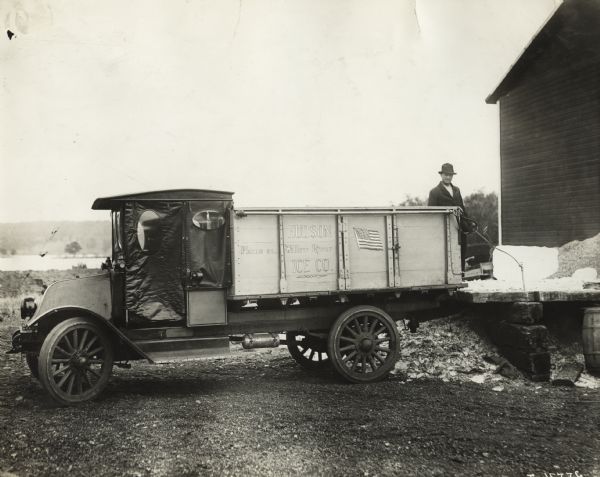 Man loading ice from loading dock with an International Model F (or 31) truck. The truck was operated by the Hudson/Willow River Ice Company. There is a lake or river in the background.