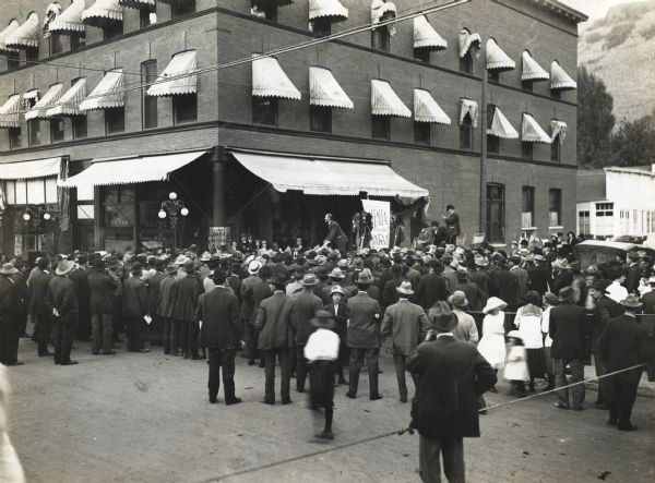 Professor Perry G. Holden demonstrates the benefits of growing alfalfa to a group of men, women, and children standing in a crossroads in a commercial area. Professor Holden stands on a raised platform in front of a brick building housing the Colfax National Bank. A photographer stands behind the professor to take a photograph of the crowd.