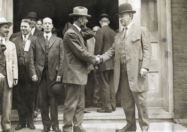Professor Perry Holden and Governor Luther Egbert Hall shake hands over a contract to grow 100,000,000 bushels of corn in Louisiana in 1913. A group of men stands in the doorway behind them looking on.
