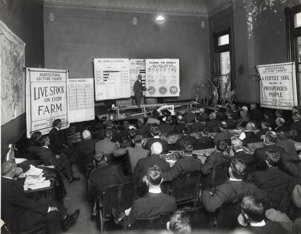 View from back of room of man standing at the front of a classroom to deliver an agricultural lecture to a group of seated men and women. There are bundles of corn on some of the tables the men are sitting at. The instructor uses several visual aid posters prepared by the International Harvester Company of New Jersey with headlines reading: "Livestock on Every Farm", "Stock Values", "All Crops of Illinois", Examine the Kernels", and "A Fertile Soil Means a Prosperous People".