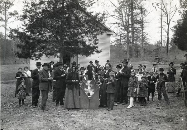 A group of men, women, and children stand outdoors in front of Shiloh Church for an outdoor meal. They are eating food laid out on a long table made out of what appears to be wooden pews or seats pushed together.