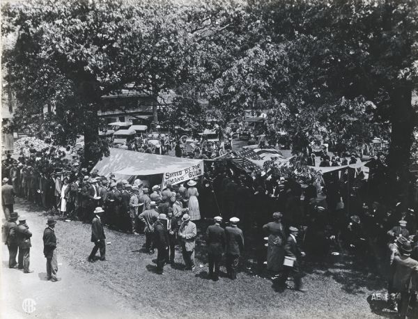 Elevated view of a crowd of people gathered around a booth on Joe Wing's farm to eat during an alfalfa presentation. A sign on the booth reads, "Served by the Woman's Guild."