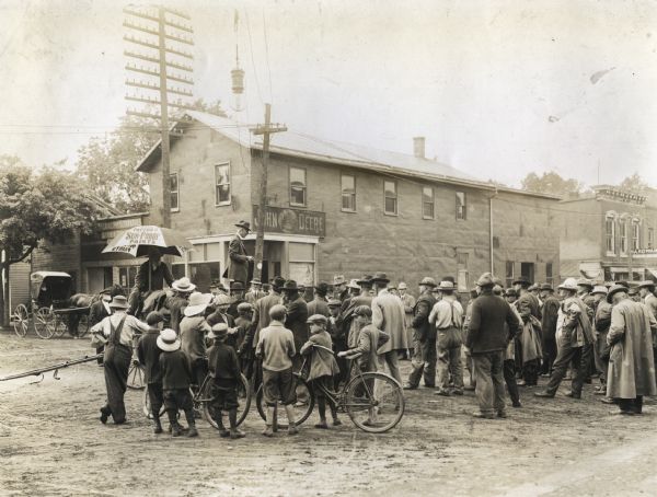 A group consisting of men and children gather around Joseph E. Wing as he stands on a platform to address the crowd on the subject of alfalfa.  The group gathers in a dirt road in front of a line of commercial buildings.