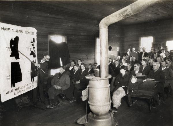 A group of men, women, and children sitting on wooden benches inside Cottage Hill School as they watch Professor P.G. Holden give a presentation on the eradication of cattle ticks. The instructor is using a pointer to direct the group's eye to a visual aid that reads: "Make Alabama Tickless. What Mississippi is Doing, Alabama Must Do." A potbelly stove stands in the foreground.