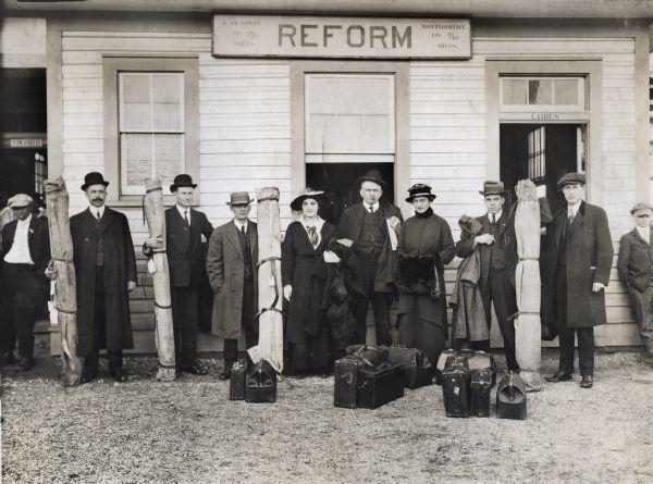 A group of speakers, six men and two women, from the Alabama Crop Diversification Campaign, stand outside a building, probably a train station, marked with a sign reading: "Reform". Some of the men hold long bundles containing educational materials, and a number of suitcases are on the ground in front of them.