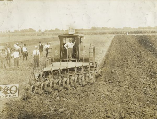 International Harvester 30-60 Mogul Tractor pulling a P&O draft plow. One man stands on the back of the tractor, and a group of men stand in the field nearby. There are stencils on the tractor, a sign on top of the tractor, and another sign on the ground near the plow.