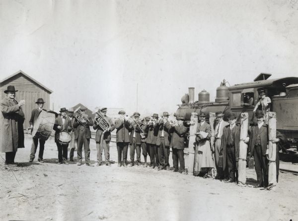 A brass band led by W.F. Casey, standing at left, welcomes four Alabama Crop Diversification Campaign speakers at a train depot. A train and several buildings are visible behind the men. Some of the men hold long bundles containing educational materials (most likely charts).