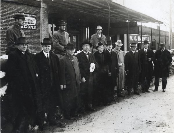 Speakers from the diversified farming program stand in front of a brick building near an awning. Each person wears a winter coat and brimmed hat, and there are cotton bales behind the group. One of the men appears to be holding some of the cotton.