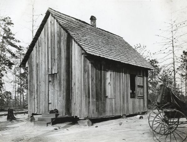 Exterior view of the New Home School. What appears to be a horse-drawn carriage stands to the right of the building. Two people can be seen sitting inside the building through an open window. A dog is lying on the ground near the window, and another dog is sitting on the steps near the front door.
