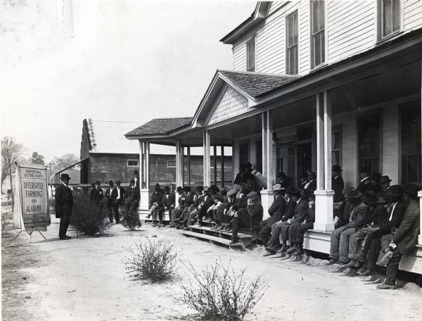 Judge Chamberlain standing near a visual aid which reads: "Agricultural Lecture Charts; Diversified Farming for Alabama," while presenting to a group of men, and some children, standing near or sitting on the porch of a hotel building.
